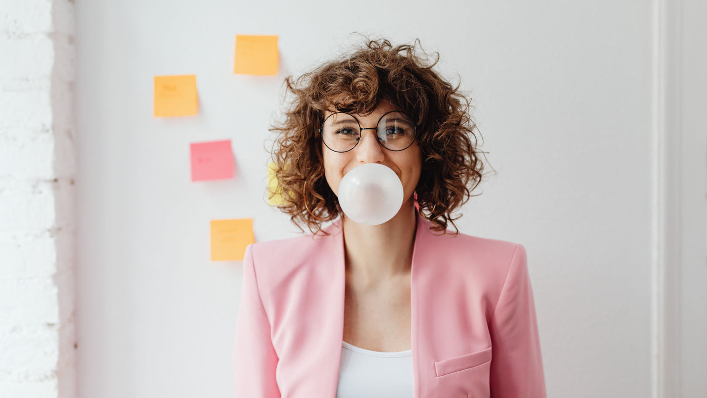 A young woman in pink blazer chewing a bubble gum with sticky notes on the wall behind her.