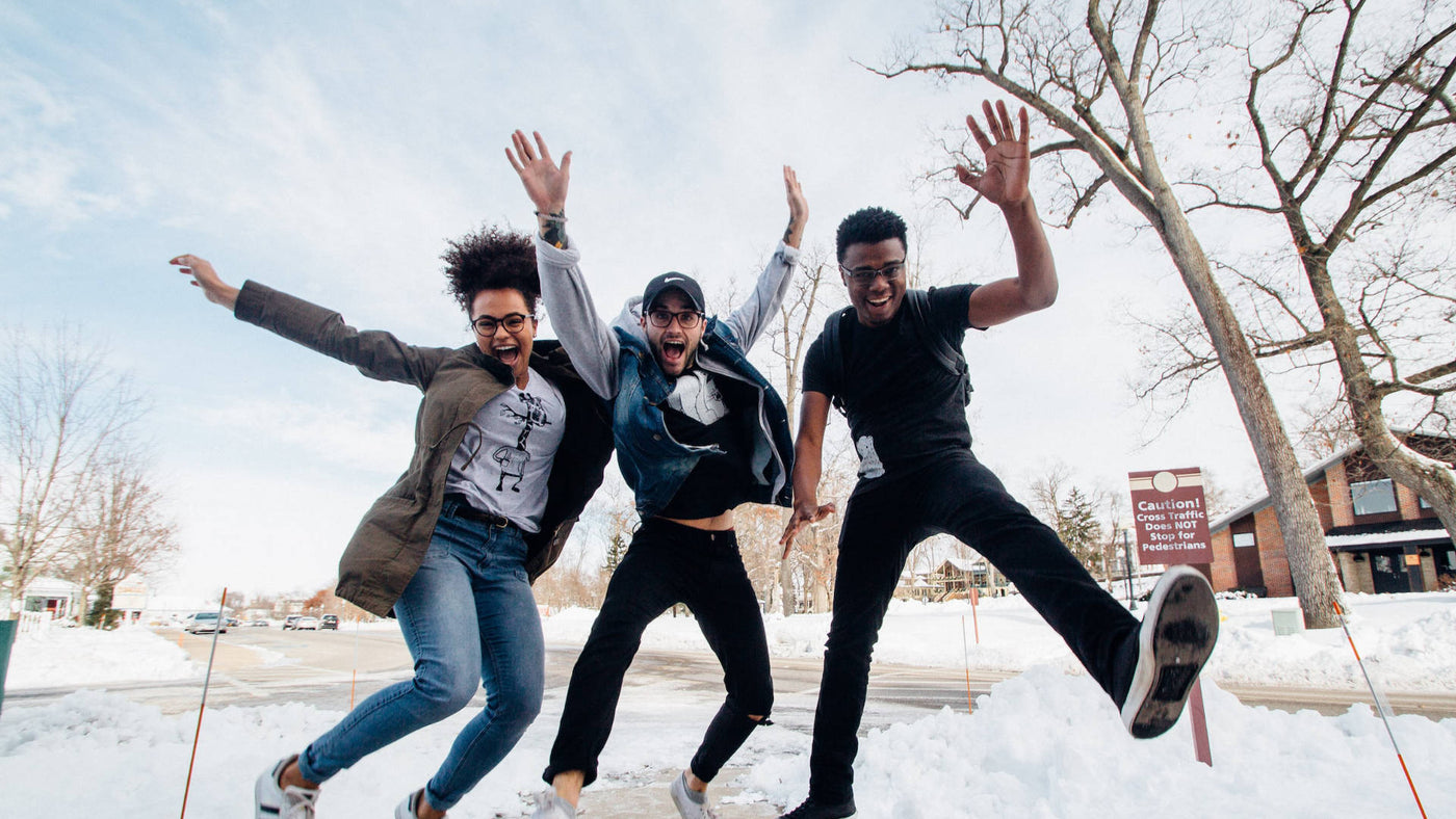 Three friends happily jumping on the ground with bare trees and snowy ground during winter.