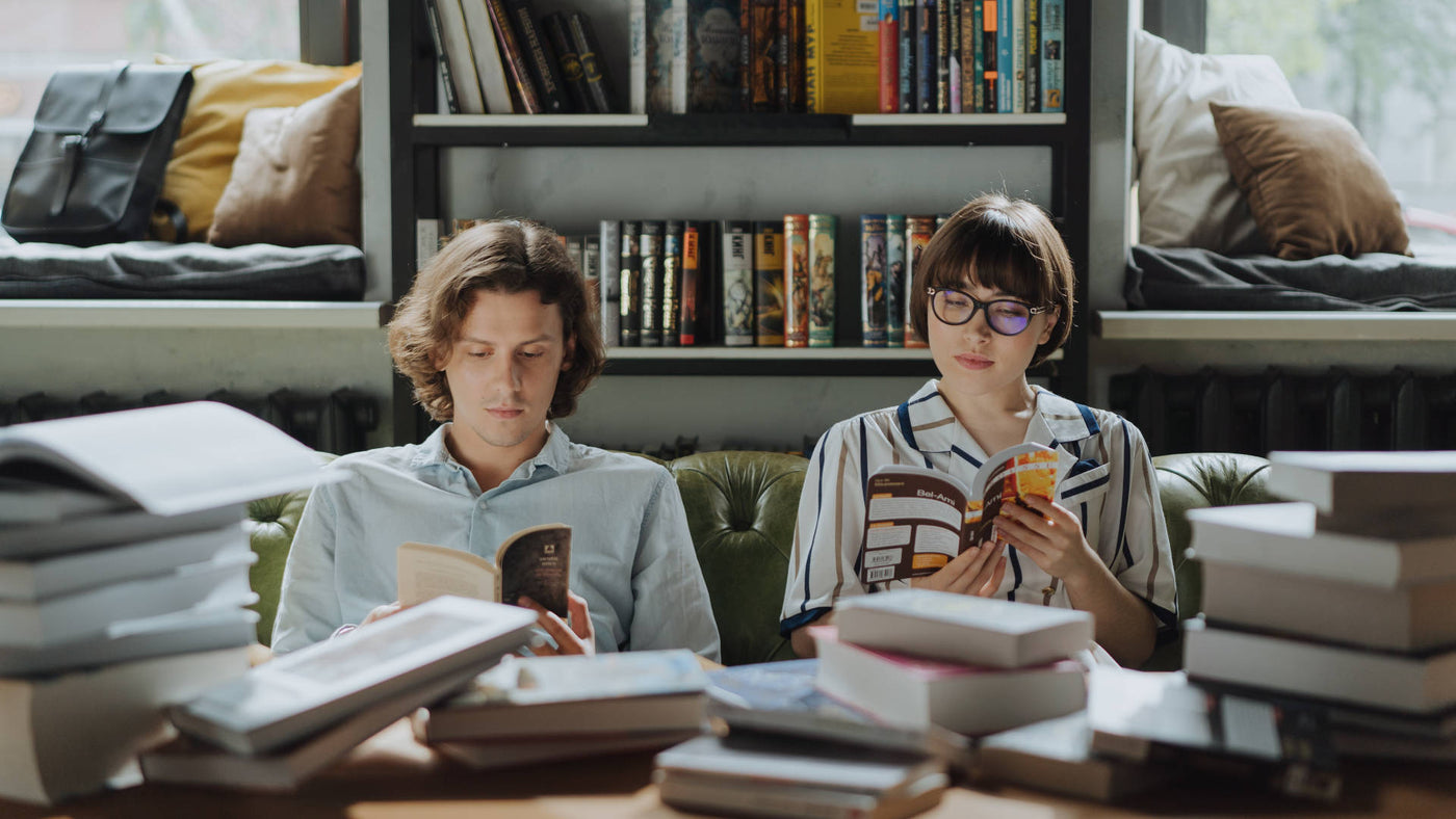 A man looking at his smartphone and a woman reading intently while both sitting on a couch with a pile of books on the table in front of them and a bookshelf at their back.