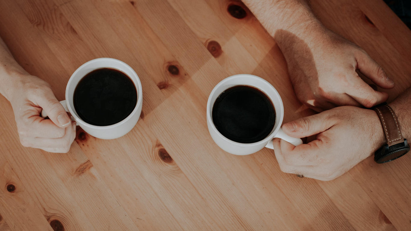 Two people holding white mugs filled with black coffee on a wooden table.