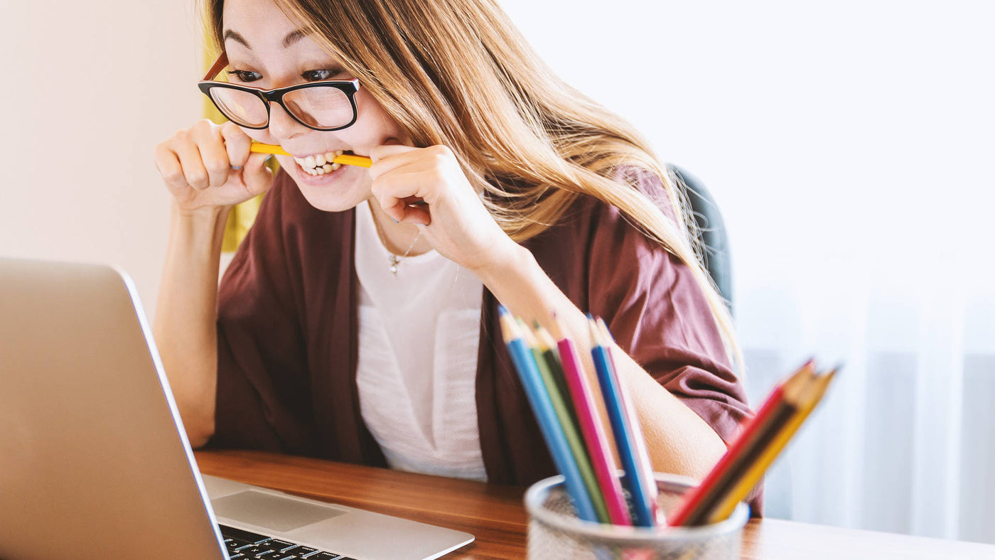 A young woman wearing reading glasses studying in front of a laptop while biting a pencil.