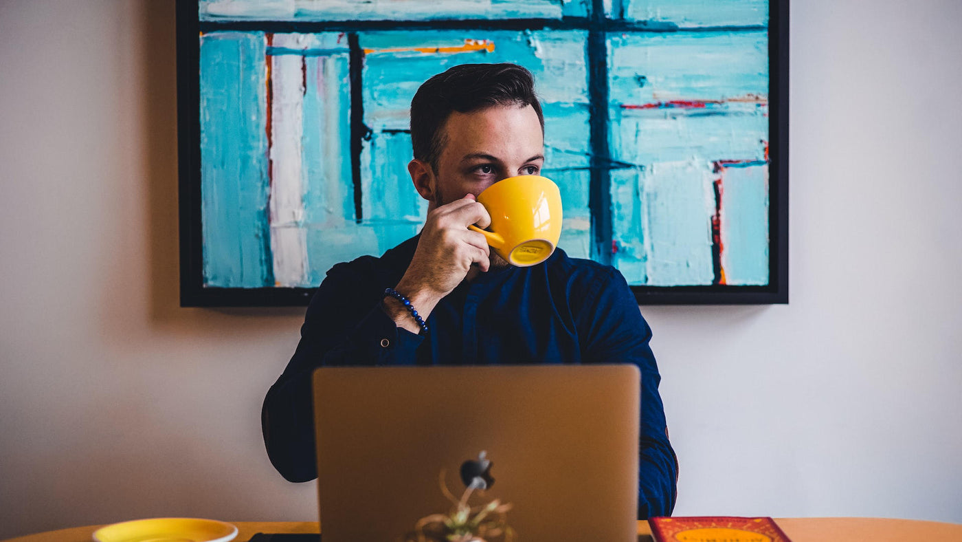 A man drinking coffee in a yellow cup while in front of a laptop.